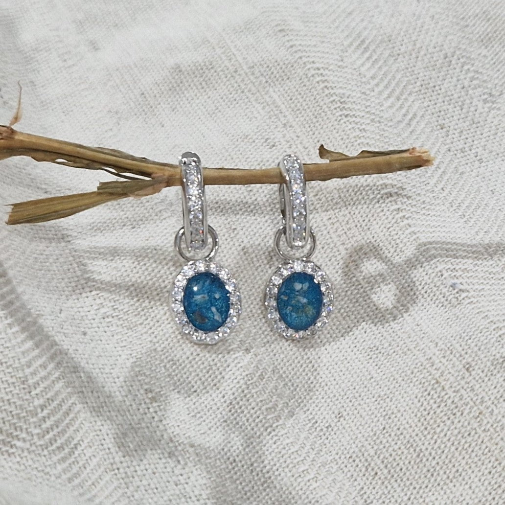 Ovaal gevormde oorhangers, 18 mm lang rondom afgewerkt met zirkonia's, aan de voorzijde zichtbaar gevuld met as of haar. Alle