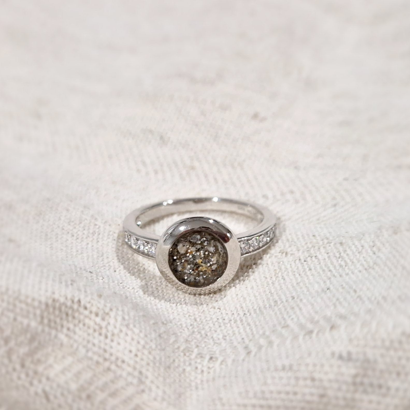 Gedenksieraad, ring 3 mm waar aan de bovenzijde zichtbaar as of haar verwerkt wordt. In de bovenste helft van de ringband zijn zirkonia's of diamanten naar keuze gezet. Alle