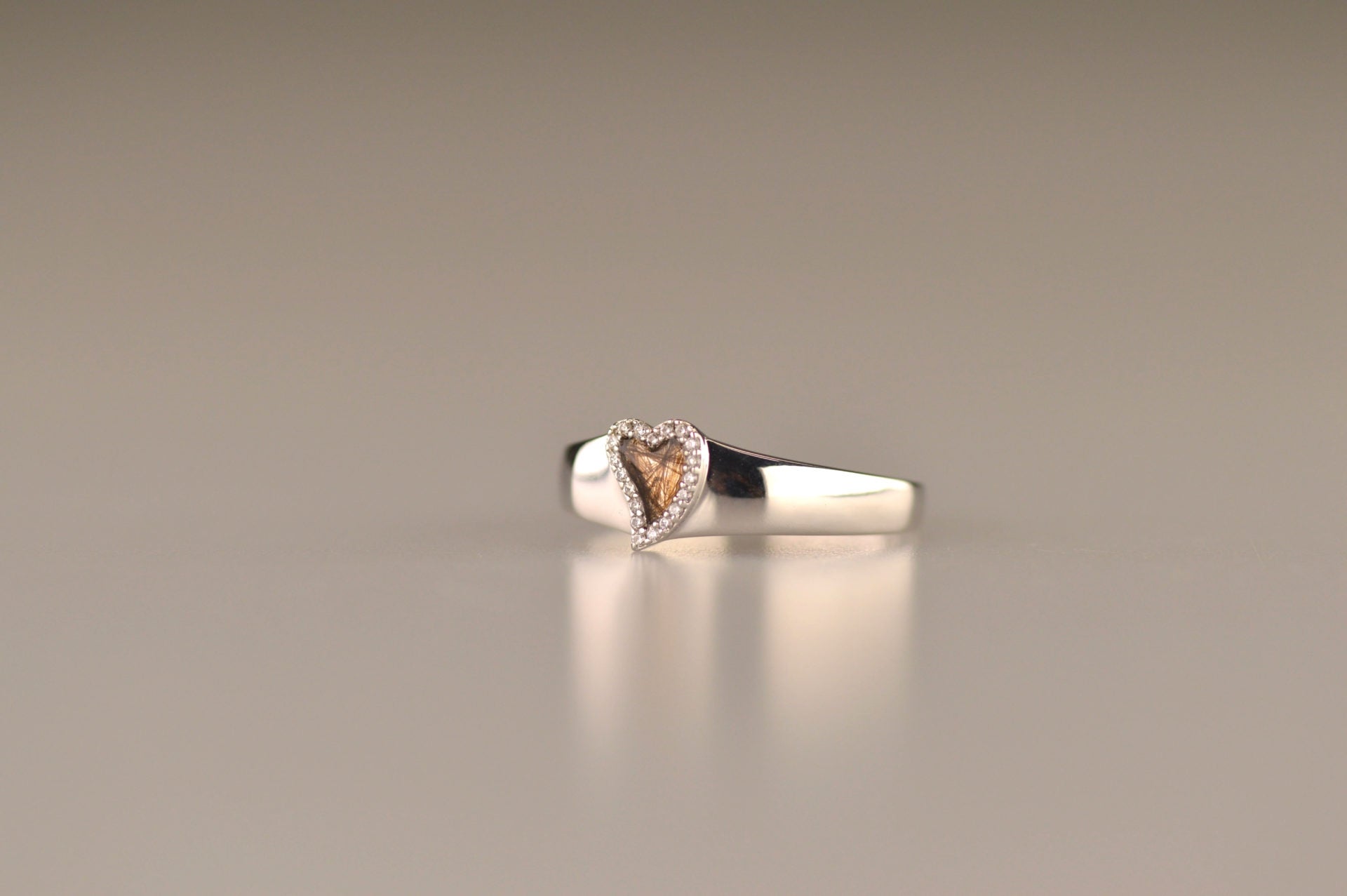 Gedenksieraad, gladde ring waar aan de bovenzijde zichtbaar as of haar (of eventueel melktandjes of moedermelk) verwerkt wordt in een hartje, rondom gezet met zirkonia's of diamanten naar keuze. alle