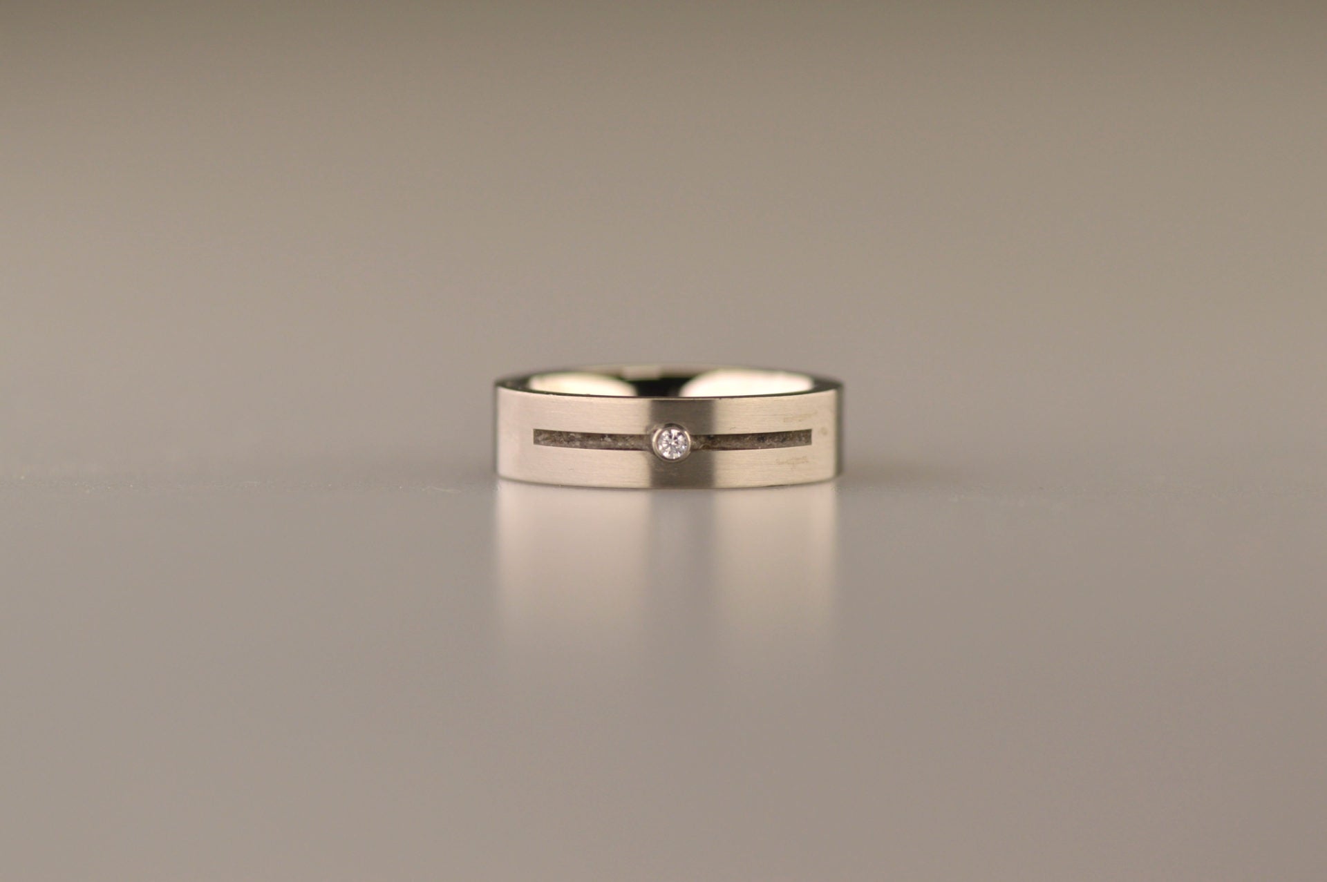 Ring als gedenksieraad 6 mm breed met één langwerpige streep, onderbroken door een zirkonia of diamant naar keuze, waardoor er twee compartimenten ontstaan, waar as of haar  in verwerkt wordt. Alle